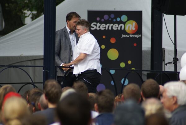 Vrouw met Flikken Maastricht-tatoeage op kuit, ongemakkelijk op podium.