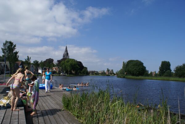 Kinderen spelen en zwemmen in de Amstel, met de St. Urbanuskerk op de achtergrond.